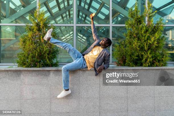 happy businessman holding mobile phone with hand raised in front of glass house - dar uma ajuda imagens e fotografias de stock