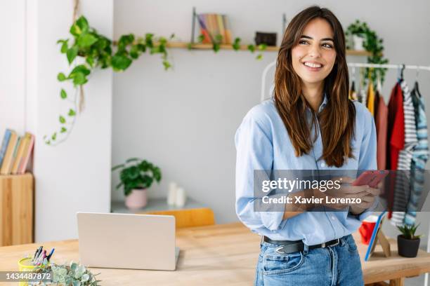 pretty small business female owner using mobile phone standing at workshop clothing store - unternehmer stock-fotos und bilder