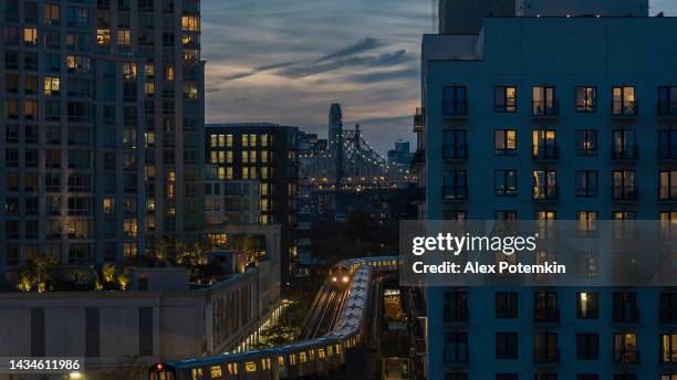 passeio de trem na linha de metrô elevada entre edifícios em long island city, queens à noite com a ponte queensboro vista atrás. - long island city - fotografias e filmes do acervo