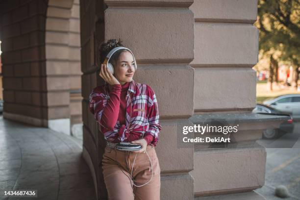 woman stands leaning against the wall and listens to music on a mini disc player, using wireless headphones - personal compact disc player 個照片及圖片檔