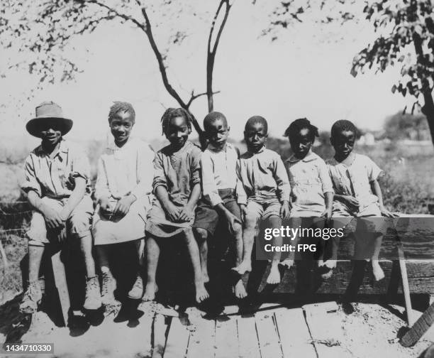 Group of children, two wearing shoes while the rest are barefoot, sit on a length of wood, United States, circa 1935.