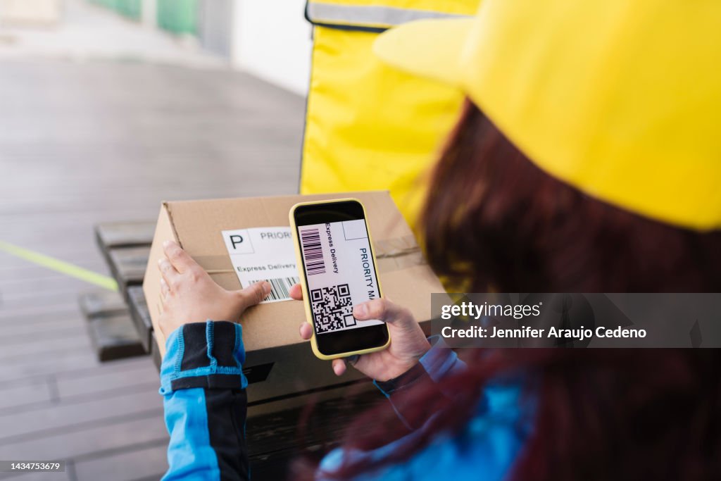 Delivery woman scans a fictitious QR code to deliver a package using a smartphone, she stands with her back to her hands getting the box and the phone close-up