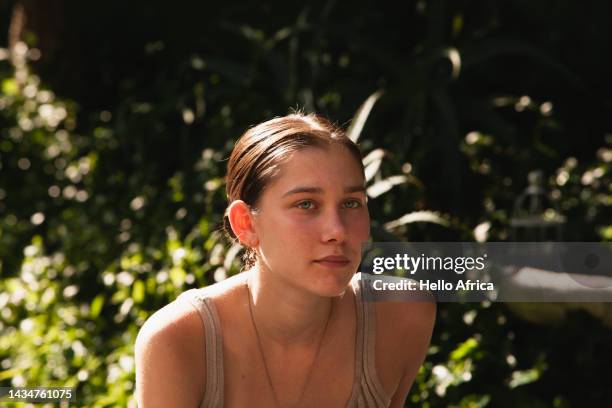 a beautiful young woman enjoying the sunshine in a leafy green garden setting, a teenage girl with long hair tied back & in a middle parting looks slightly away from camera with a pensive & absorbed facial expression - female looking away from camera serious thinking outside natural stock pictures, royalty-free photos & images