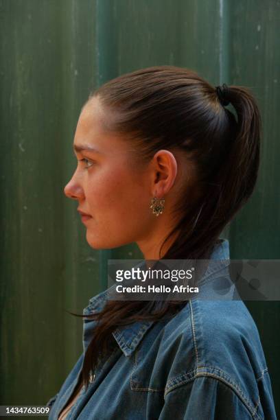 profile view of a beautiful young woman on a green background, a teenage girl with long brown hair tied back in a ponytail & wearing a blue denim shirt & ornate hanging earring looks directly ahead of herself in profile with a slight smile on her face - drop earring stock-fotos und bilder