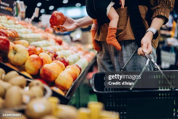 cropped shot of young asian mother holding a shopping basket, grocery shopping with her baby daughter in supermarket. mother carrying her baby girl in carrier and choosing fresh organic apples. fruits and vegetables shopping. healthy eating lifestyle - apple products stock pictures, royalty-free photos & images
