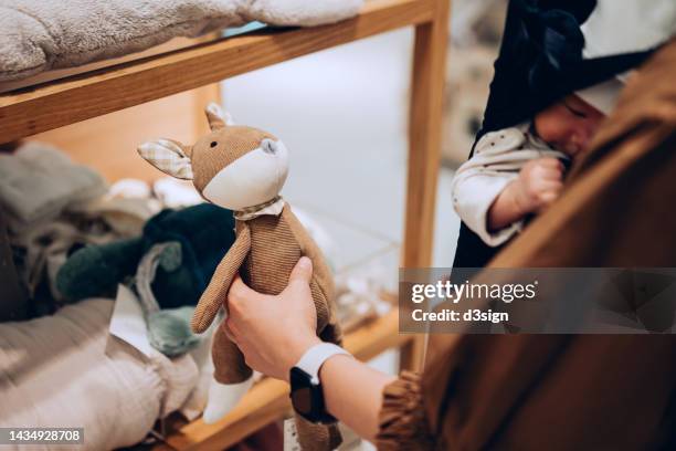 cropped shot of young asian mother carrying her baby girl in carrier, shopping for soft toys and baby essentials in a boutique. mother shopping for kids products and essentials in a store - baby products stock pictures, royalty-free photos & images