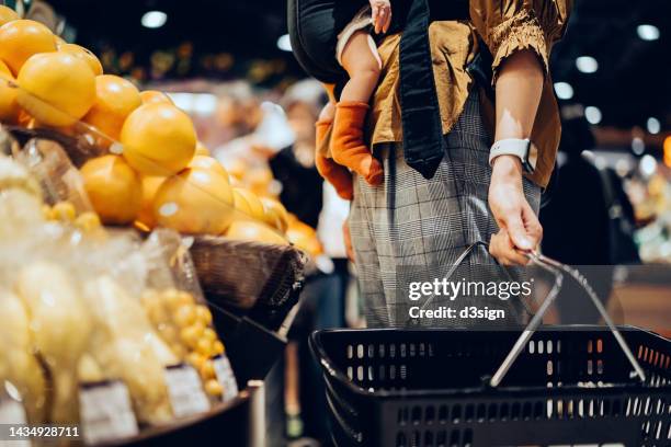 cropped shot of young asian mother holding a shopping basket, grocery shopping with her baby daughter in supermarket. mother carrying her baby girl in carrier and choosing fresh organic produce. fruits and vegetables shopping. healthy eating lifestyle - homegrown produce ストックフォトと画像