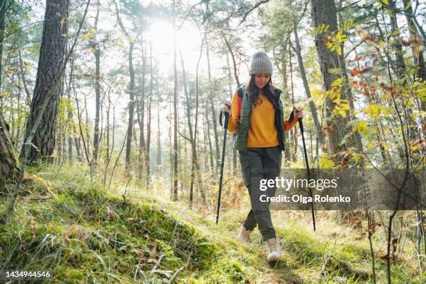 full length portrait of young woman with backpack and nordic walking poles enjoying hiking in forest lit by sunlight - nordic walking stock pictures, royalty-free photos & images