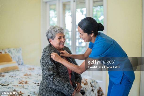 老人ホームの寝室で年配の女性がシャツを着るのを手伝うミッドアダルト看護師 - 老年医学 ストックフォトと画像