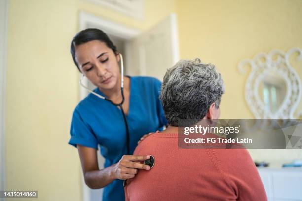 krankenschwester im mittleren erwachsenenalter, die die lungen einer älteren frau mit einem stethoskop im schlafzimmer eines pflegeheims untersucht - adult patient with doctor and stethoscope stock-fotos und bilder