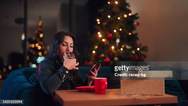 jeune femme mangeant des aliments sucrés et appréciant d’utiliser son téléphone intelligent tout en se relaxant dans le salon à la maison pendant noël - red telephone box photos et images de collection