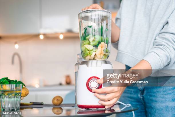 a young woman whips fruits and vegetables in a blender to make a smoothie. - blended drink stock pictures, royalty-free photos & images