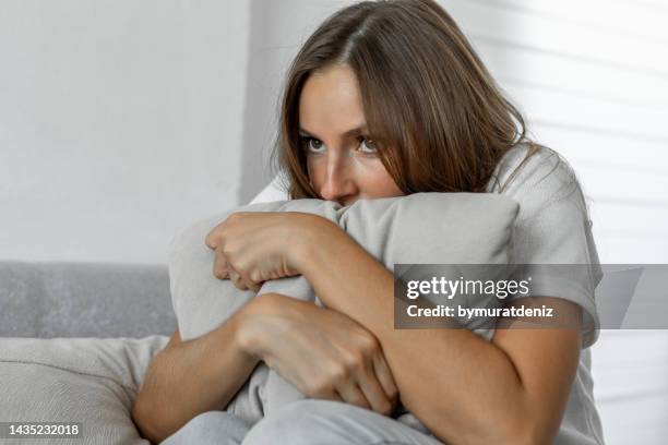 scared woman at home embracing pillow sitting on a sofa - fobia imagens e fotografias de stock