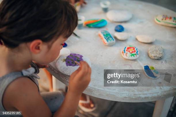 the girl paints on white stones. on the table in front of her are stones that she has already decorated - rock art stock pictures, royalty-free photos & images