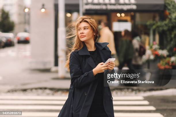 a young confident business woman in a coat looks into a mobile phone prints an online message using wireless technology crosses the road on the street in the city of outdoor. a beautiful serious girl student freelancer influencer and millennial - russian business woman stock-fotos und bilder