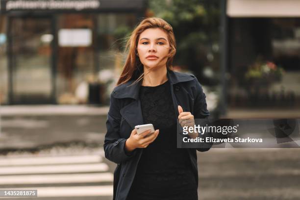 a young confident business woman in a coat looks into a mobile phone prints an online message using wireless technology crosses the road on the street in the city of outdoor. a beautiful serious girl student freelancer influencer and millennial - russian business woman stock-fotos und bilder