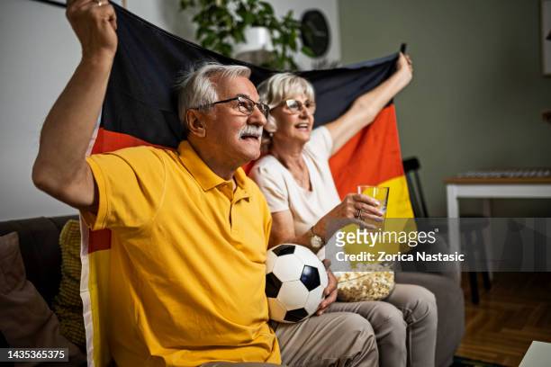 seniorenpaar schaut fußballspiel im wohnzimmer mit deutscher flagge im fernsehen - fußball fan oma stock-fotos und bilder