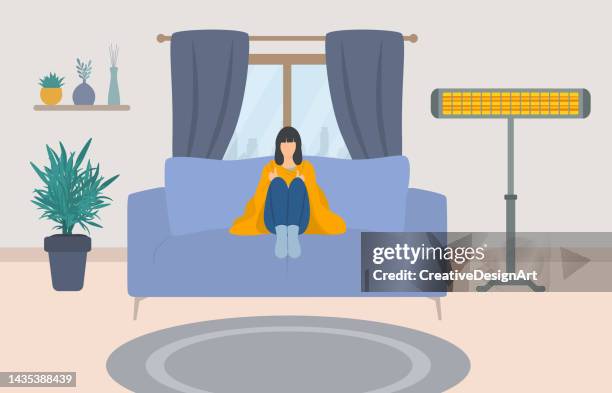 young woman wrapped in blanket, sitting on couch.electric heater in living room - cold house stock illustrations