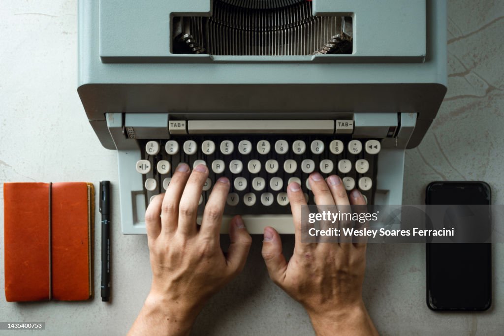 Flat lay image of human man hands typing old typewriter on a business office desk with leather notepad and cellphone
