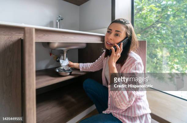 woman having a leak in her kitchen sink and calling the plumber - emergencies and disasters imagens e fotografias de stock