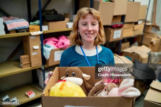 cheerful young woman at a charitable foundation holding a box full of toys to donate - charitable foundation stockfoto's en -beelden