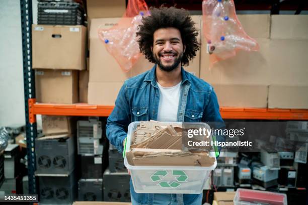 black happy young man working at a recycling centre holding a container with cardboard facing camera smiling - circulaire economie stockfoto's en -beelden