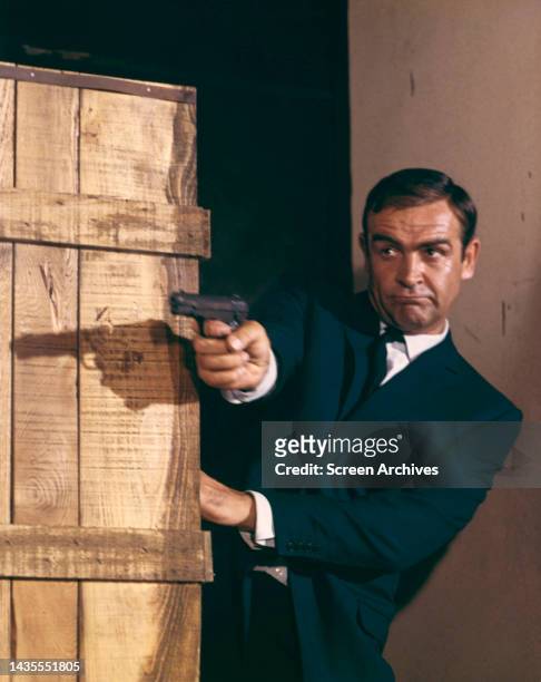 Sean Connery as James Bond 007 firing pistol behind crates from Goldfinger 1964.