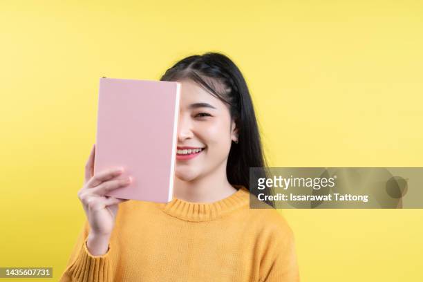 portrait of a pretty young girl hiding behind an open book and looking away isolated over yellow background - open book stock-fotos und bilder