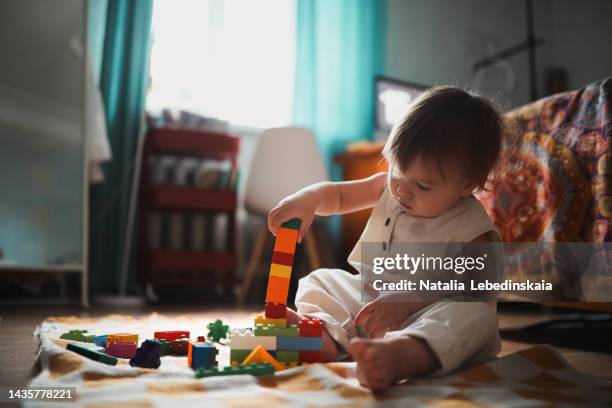 kid in linen jumpsuit sits alone and plays with educational toys at home. toddler boy 1 year old builds tower of plastic blocks on floor of apartment. real interior. - plastic toy stock pictures, royalty-free photos & images