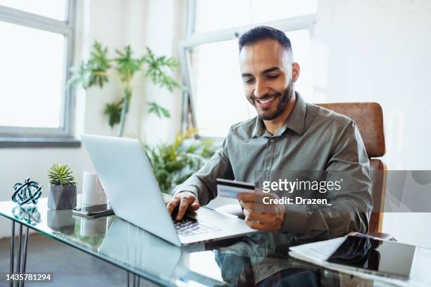 mid adult smiling latin businessman in office, using credit card to pay online - betalen stockfoto's en -beelden