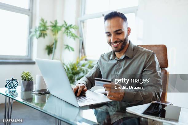 mid adult smiling latin businessman in office, using credit card to pay online - corporate business transaction stock pictures, royalty-free photos & images