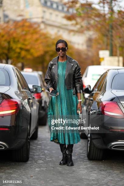 Guest wears black and green sunglasses from Prada, gold earrings, a green pleated long dress, a black shiny leather zipper biker jacket, a black...