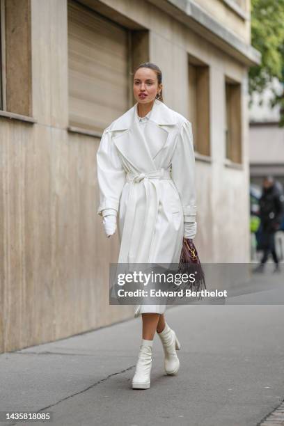 Guest wears silver earrings, a white shirt, a white shiny leather belted long trench coat, a burgundy shiny leather fringed handbag, white latte...