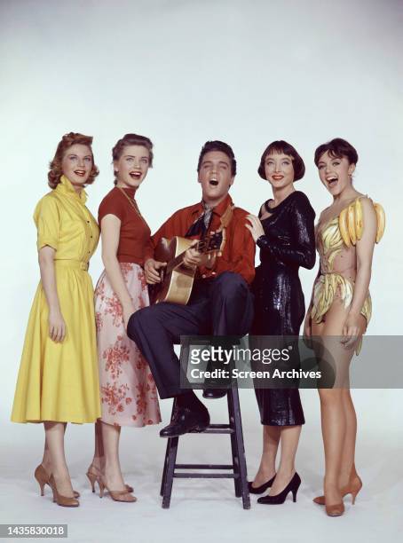 Elvis Presley vivid color studio publicity pose of "The King" sitting on stool as he plays guitar with Carolyn Jones, Delores Hart, Liliane...