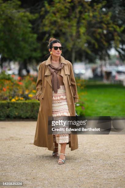 Guest wears black sunglasses from Ray Ban, gold large earrings, a brown wool pullover, a brown t-shirt, a beige long trench coat, a white latte and...
