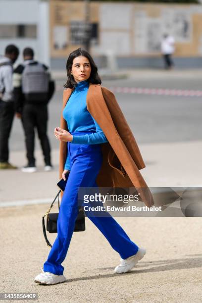 Guest wears a blue turtleneck wool pullover, royal blue flared suit pants, a brown long wool coat, a black matte leather handbag, white leather and...
