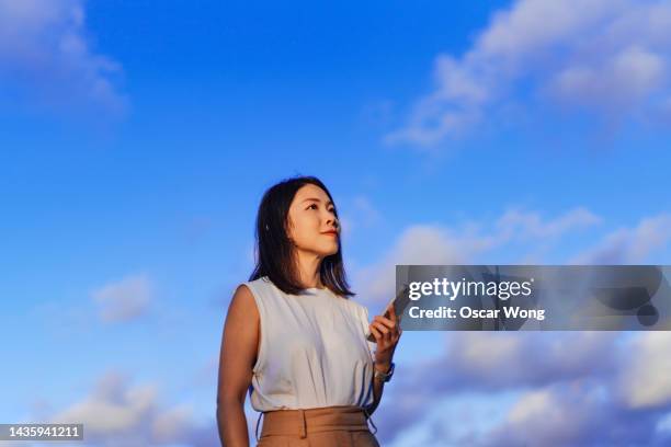 confident young asian woman looking up while using smartphone, standing against blue sky - japanese bussiness woman looking up imagens e fotografias de stock