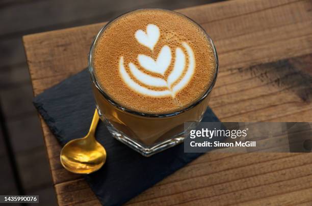 flat white coffee with heart-shaped froth art in a drinking glass on a slate coaster with a brass teaspoon on a wooden outdoor cafe table - color crema stockfoto's en -beelden