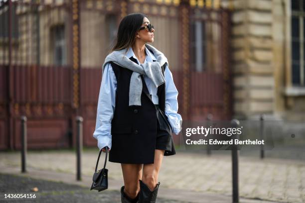 Angela Gonzalez wears black sunglasses, gold earrings, a white oversized shirt, a pale gray wool pullover knot at the waist, a black sleeveless /...