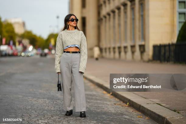 Angela Gonzalez wears black sunglasses from Celine, gold earrings, a pale gray embossed pattern cropped wool pullover, a pale blue cropped shirt, a...