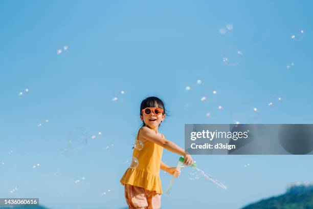 happy little asian girl with sunglasses having fun playing and blowing soap bubbles outdoors on a beautiful sunny day against blue sky - child blowing bubbles stock-fotos und bilder