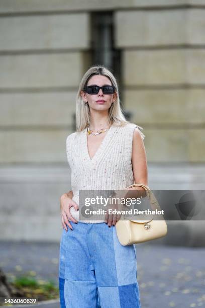 Natalia Verza wears black sunglasses, silver earrings, a white pearls / yellow necklace, a white latte wool V-neck / sleeveless pullover, a brown...