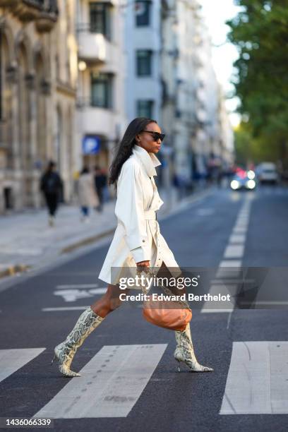 Emilie Joseph wears black sunglasses, diamonds earrings, a white minimalistic belted trench coat from Isabel Marant, a brown matte leather puffy...