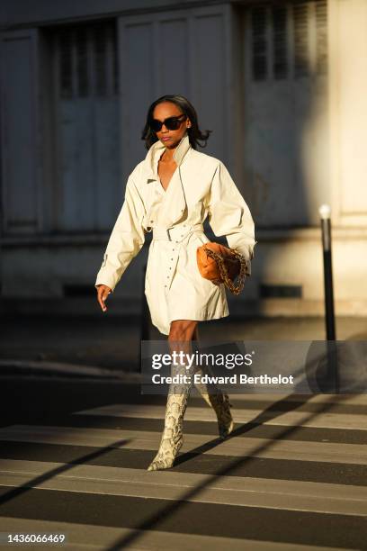 Emilie Joseph wears black sunglasses, diamonds earrings, a white minimalistic belted trench coat from Isabel Marant, a brown matte leather puffy...