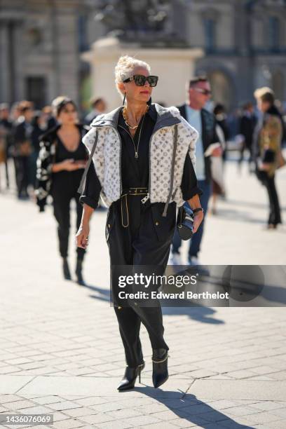 Guest wears black sunglasses from Louis Vuitton, a gold large chain necklace from Louis Vuitton, a black zipper neck shirt, a white with black LV...