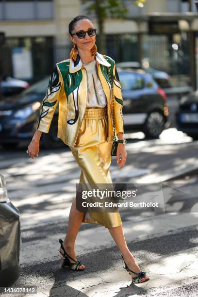 Guest wears black sunglasses, gold large leather pendant earrings, a gold shiny t-shirt, a gold shiny leather midi skirt, a gold shiny leather with...