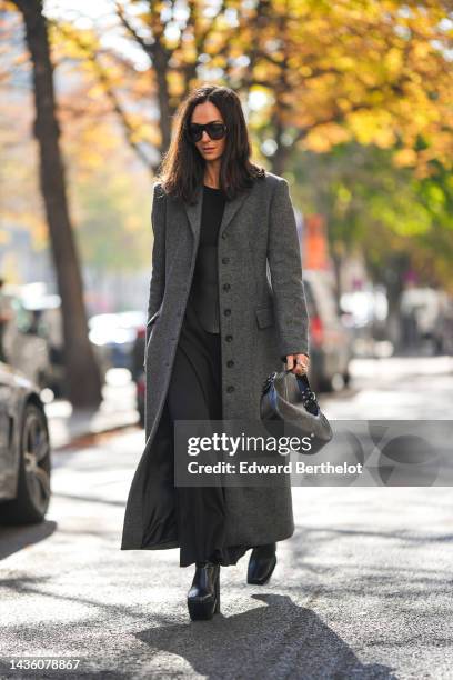Guest wears black sunglasses, a black ribbed long dress, a gray long wool coat, a black shiny leather nailed / studded handbag, black platform wood /...
