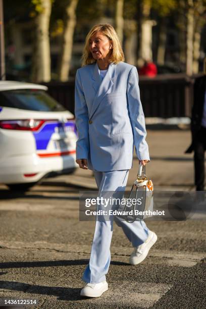 Guest wears a white t-shirt, a pale blue blazer jacket, matching pale blue suit pants, a white with brown / beige fur cow print pattern handbag,...