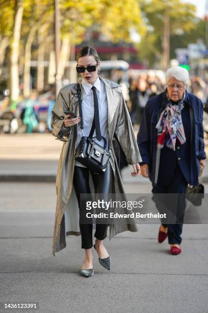Guest wears black square sunglasses, silver earrings, a white shirt, a black tie, a silver shiny leather long coat, a black shiny leather nailed /...