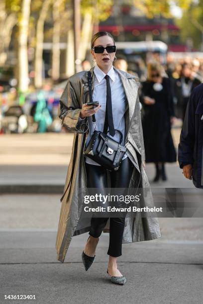 Guest wears black square sunglasses, silver earrings, a white shirt, a black tie, a silver shiny leather long coat, a black shiny leather nailed /...